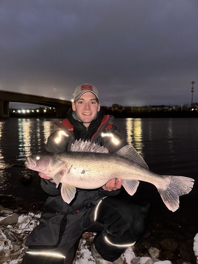 Phillip kneeling on a snowy riverbank at dusk, holding a large walleye